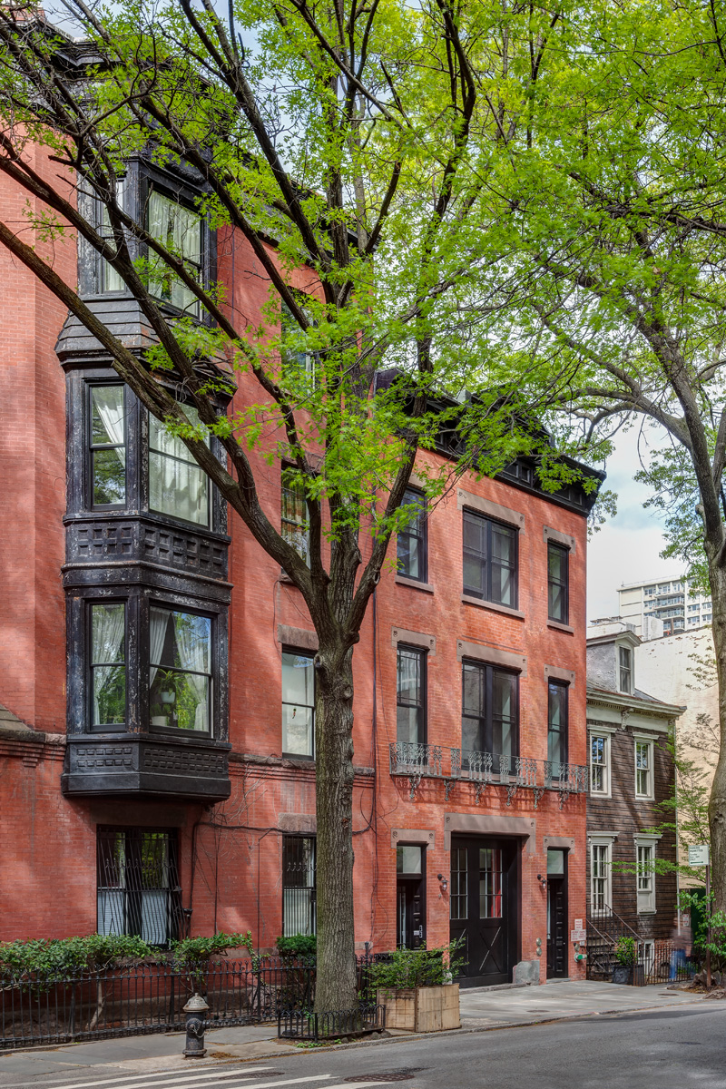 Front facade of a three-story brick rowhouse with black doors, a black cornice, and brownstone accents. A large tree sits on the sidewalk in front of it.