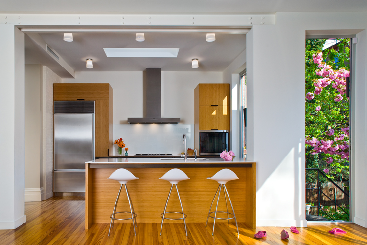 Kitchen with stainless steel appliances, three white barstools in front of the counter, flush mount lighting, a skylight, and large windows to the rear yard.