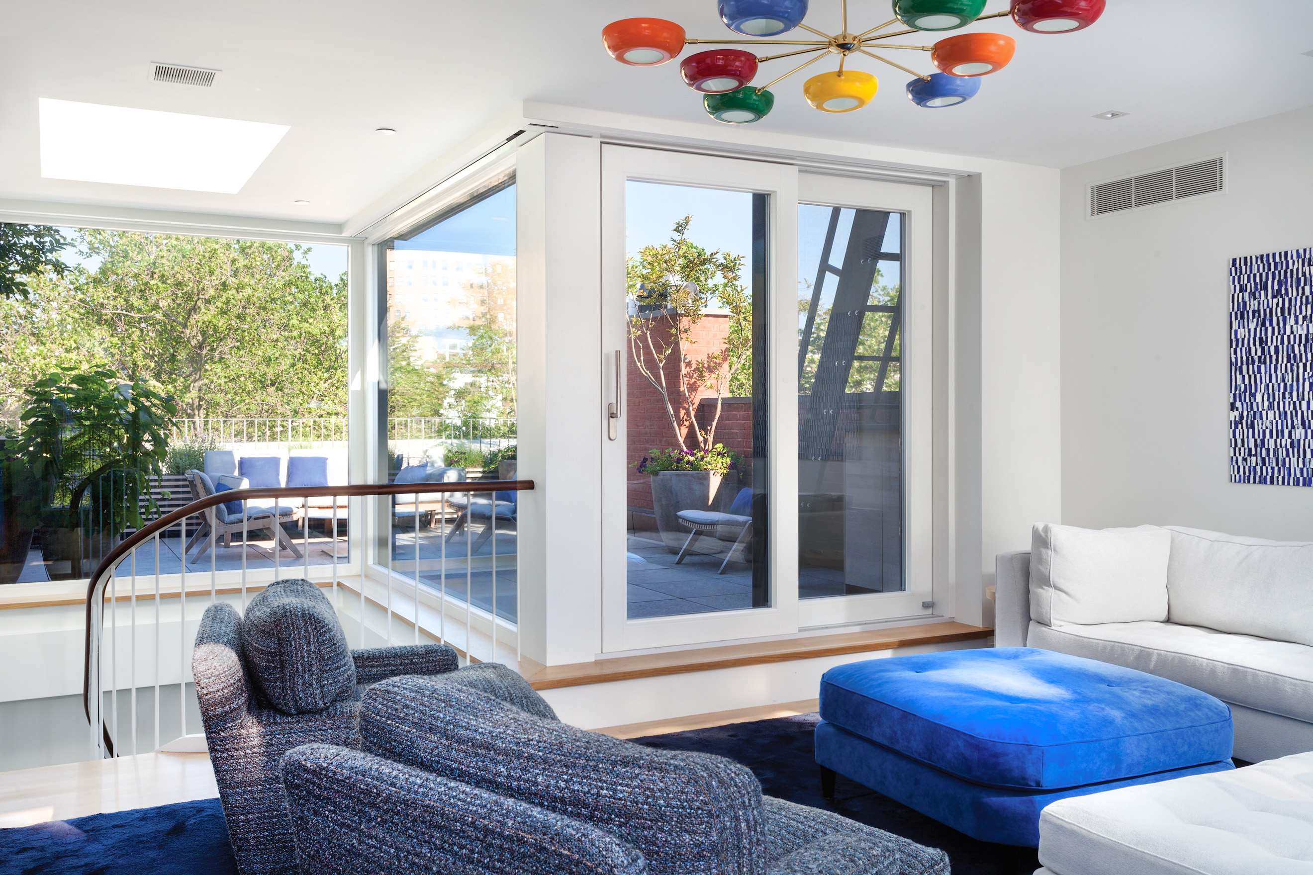 Penthouse in a Brooklyn Heights townhouse with blue carpet, painting, ottoman, and armchairs. A colorful light fixture hangs off the ceiling, and the rooftop deck can be seen through the large windows and glass door.