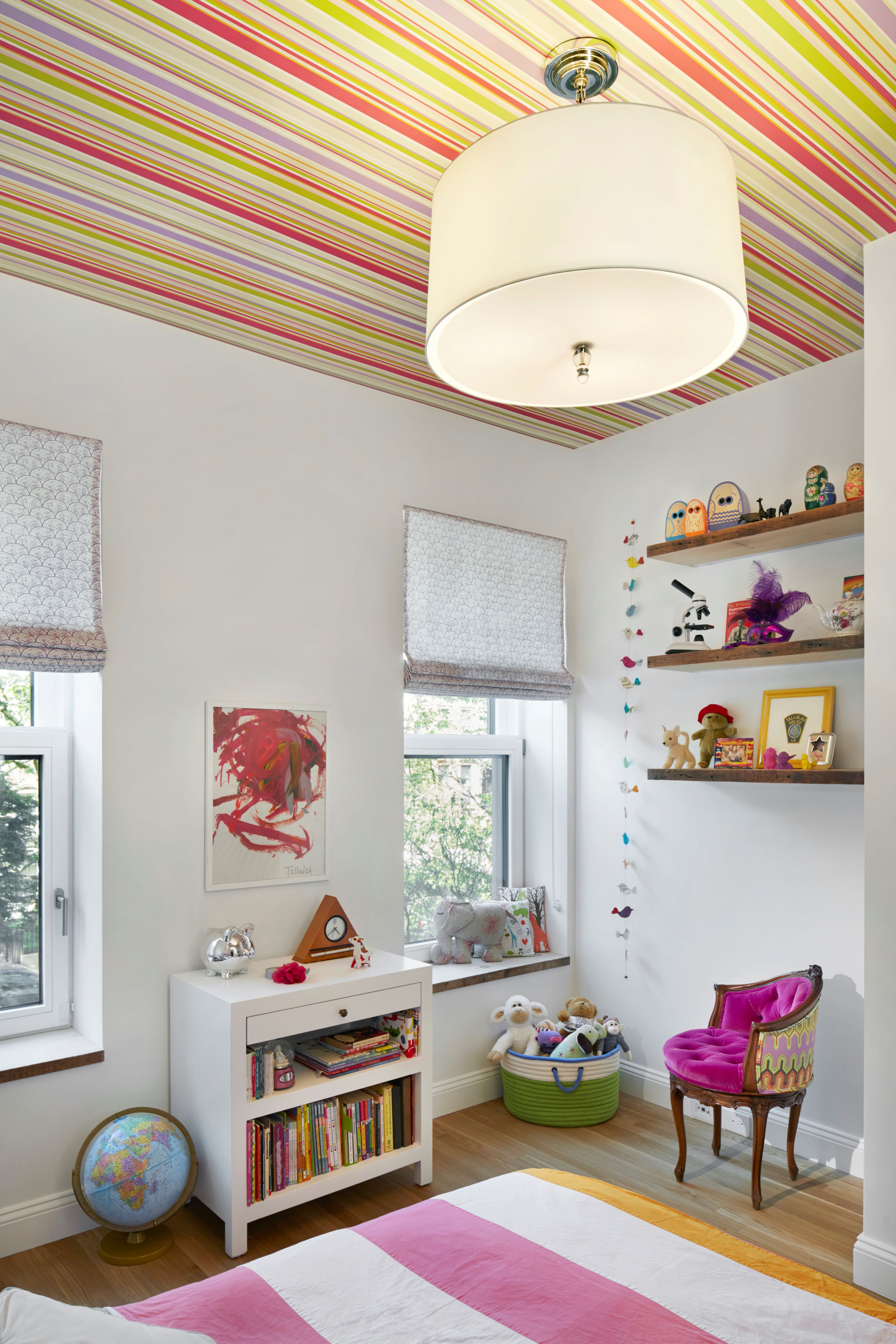 Corner of a child's bedroom with striped wallpaper on the ceiling, suspended bookshelves, Roman shades, and a white and pink striped comforter at the bottom of the frame.