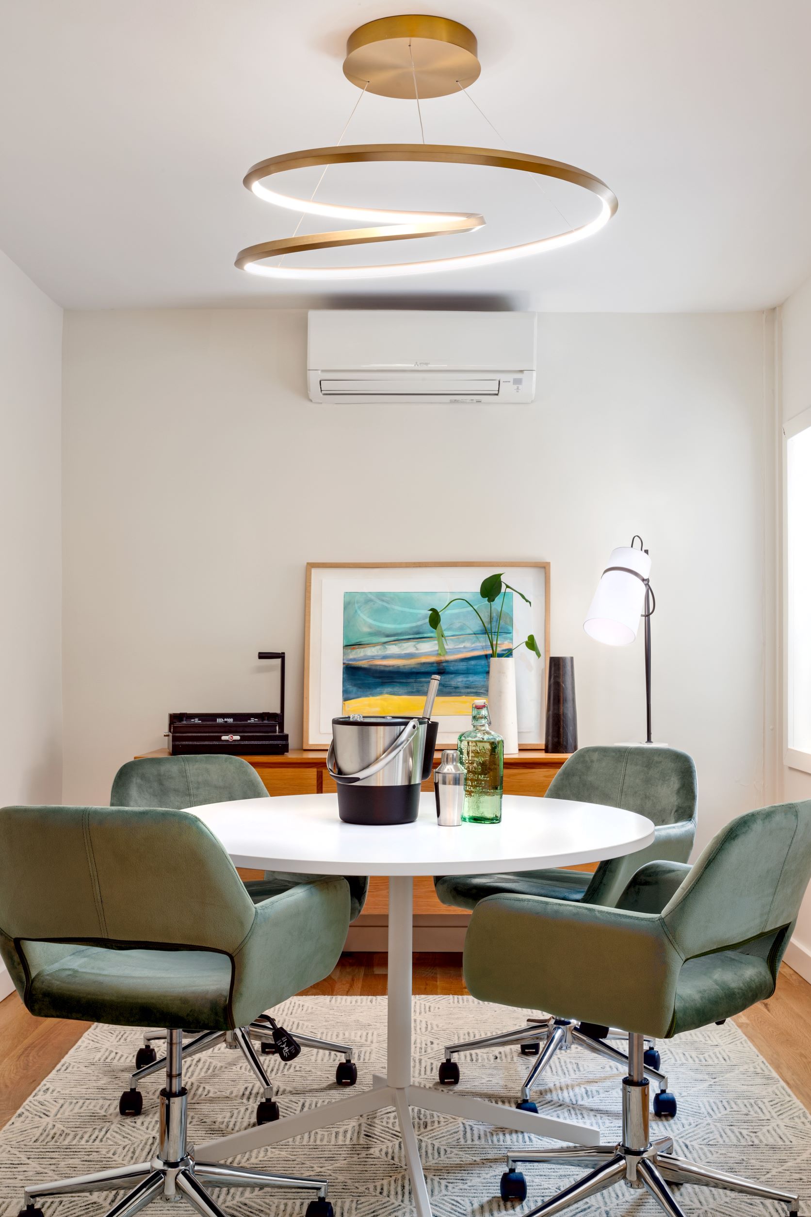 Office space with an abstract gold chandelier hanging above a round conference table with four green velvet chairs. Bar accoutrements sit atop the conference table, and an area rug sits below it.
