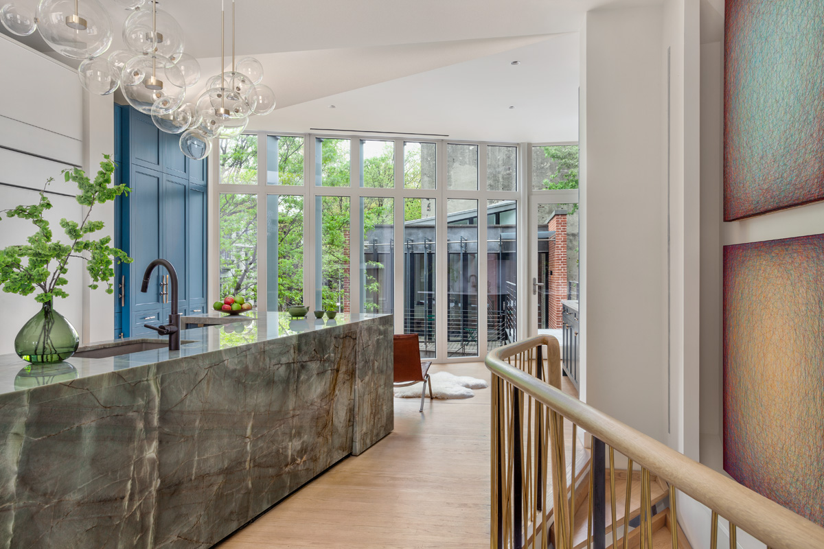 View from kitchen, across a courtyard, and into an exercise studio. A large marble island is in the front left foreground, and wood staircase leading downstairs in the front right foreground