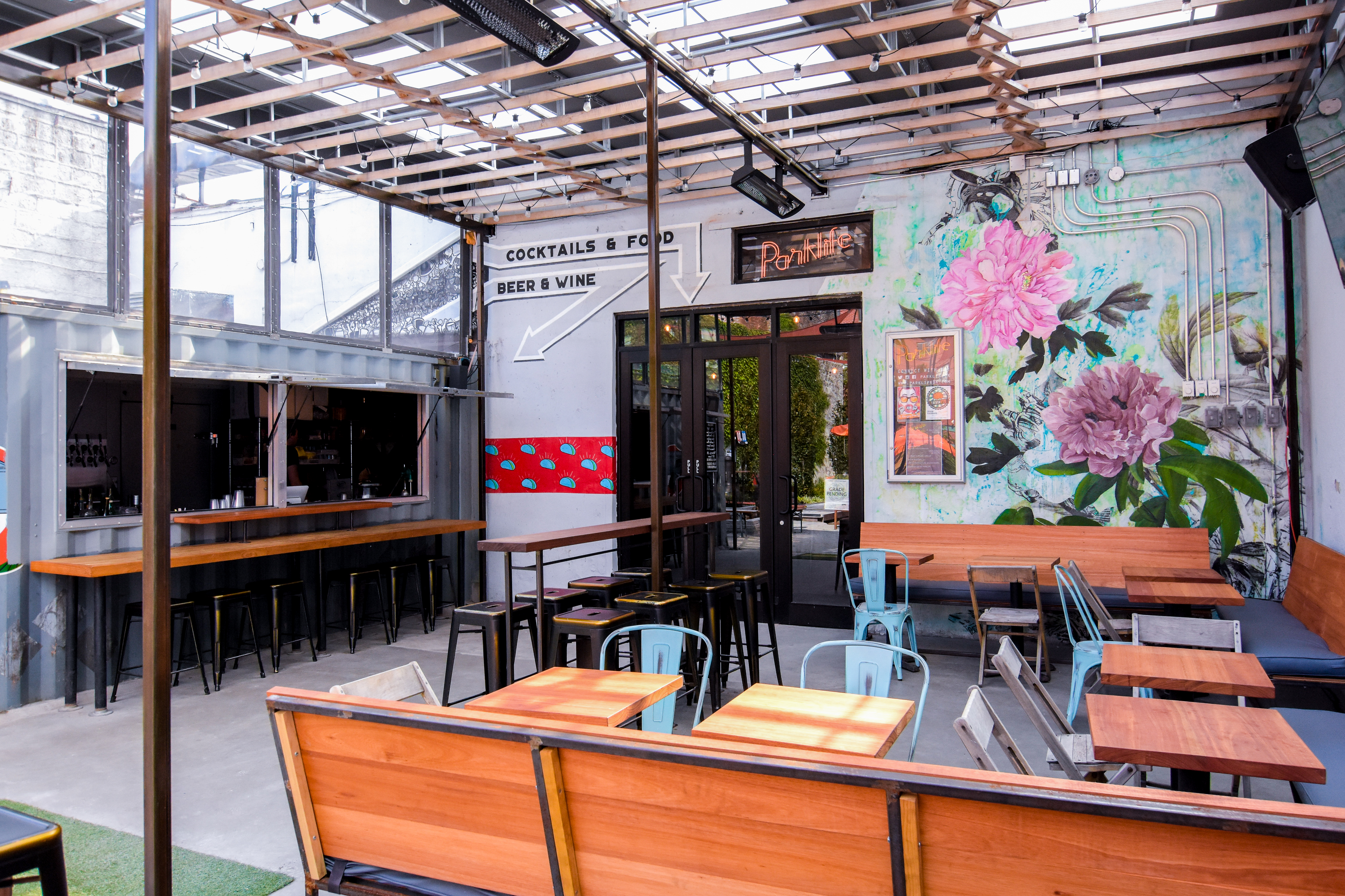 Entrance to Parklife with a pergola, wood tables and bars, blue chairs, and black metal barstools.