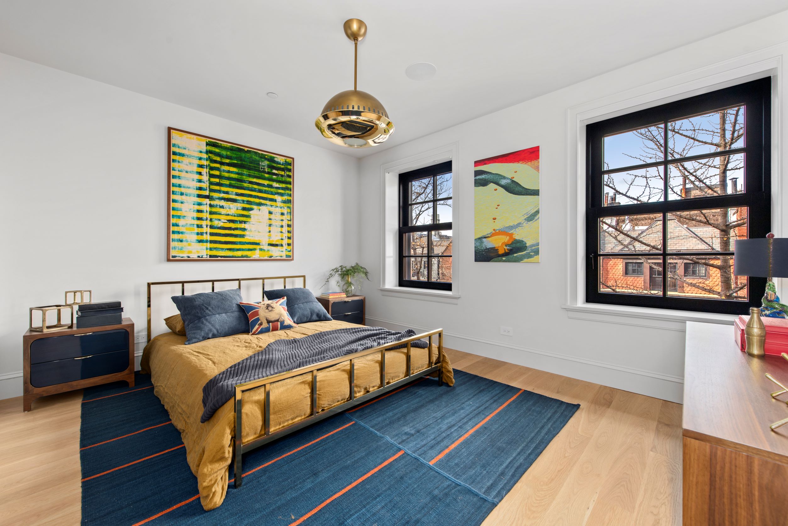 Bedroom with a large gold chandelier, wood floors, blue area rug underneath a bed with a mustard-yellow comforter, and wood dresser.