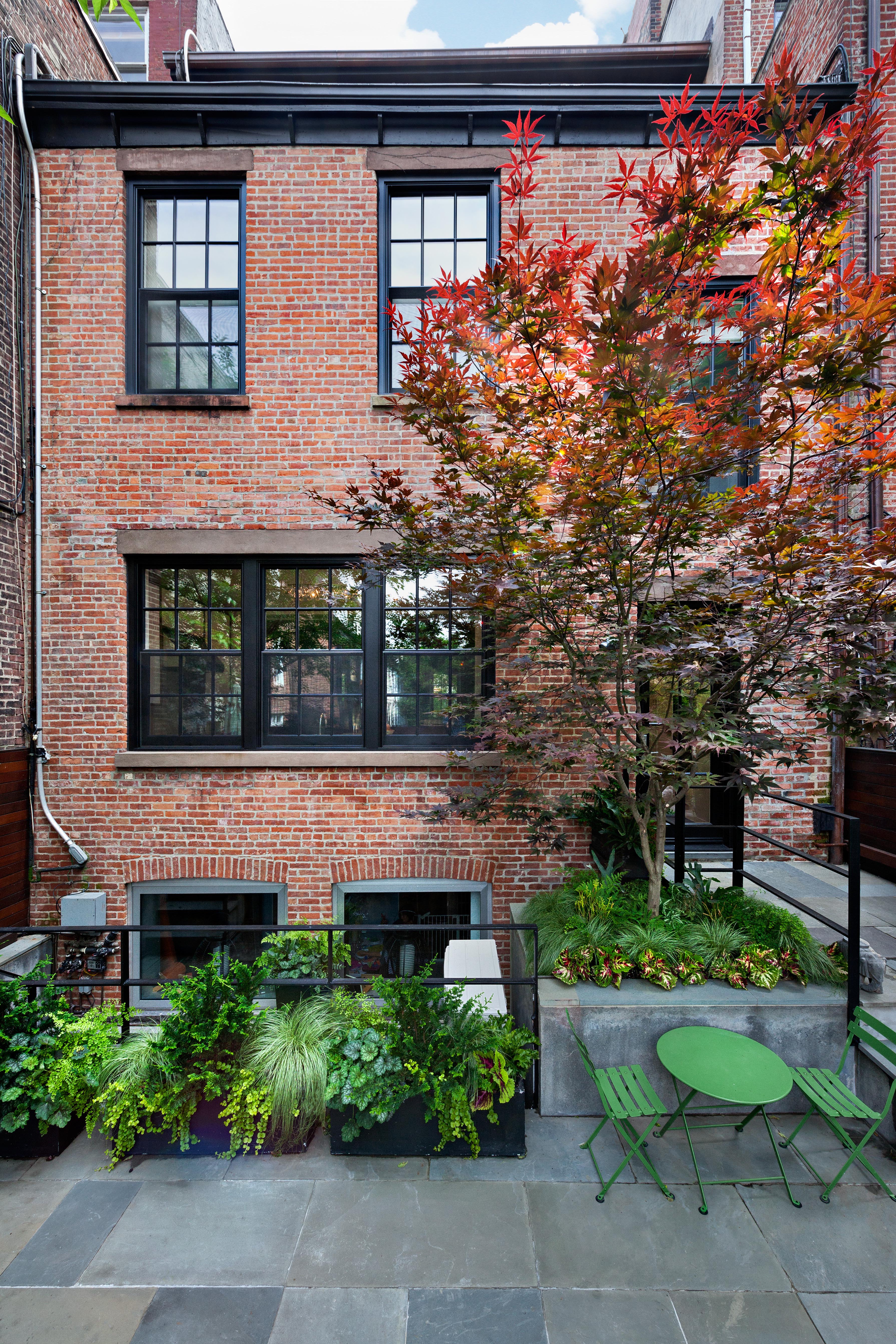 Rear façade of a brick house with a red tree, bluestone pavers, and green table and chairs